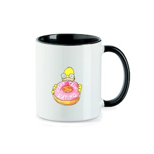 Könnte beinhalten: Weiße Keramiktasse mit schwarzem Rand und Henkel. Die Tasse zeigt ein Cartoon-Bild eines gelben Charakters mit einem Donut im Mund. Der Donut ist rosa mit weißem Zuckerguss und dem Text "Can't Talk Eating".