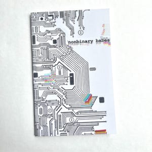 以下が含まれることがあります： 黒い回路基板のデザインが施された白い小冊子。「nonbinary babes」の文字が前面に印刷されています。回路基板にはカラフルな虹色のデザインが見えます。