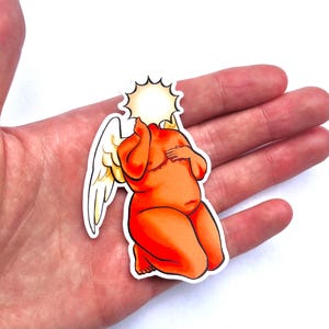 Puede incluir: Una pegatina que representa un querubín naranja estilizado con una cabeza de sol y alas de ángel. El querubín está arrodillado, con una mano en el pecho y la otra cerca de la cara. La pegatina tiene un borde blanco.