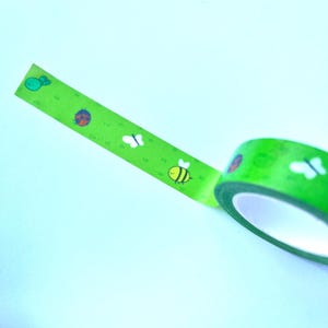 May include: Green washi tape with a repeating pattern of a ladybug, butterfly, and bee. The tape is rolled up and partially unrolled, showing the pattern.