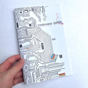 Puede incluir: Un cuaderno blanco con un diseño de placa de circuito negro y el texto "nonbinary babes". El cuaderno está sostenido por una mano.