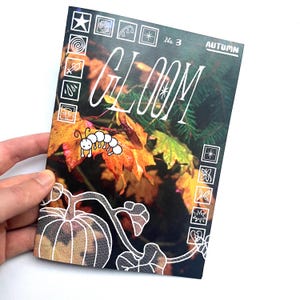 Può includere: Una copertina di rivista con il titolo "GLOOM" in scrittura bianca. La copertina presenta una scena di fogliame autunnale con foglie arancioni e gialle, un bruco dei cartoni animati e la parola "AUTUMN". Una mano tiene la rivista.