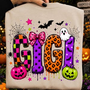 May include: A beige sweatshirt featuring the text GIGI in large, colorful letters. The letters are decorated with checkered, polka dot, and leopard patterns. The design includes a pink bow, a ghost, pumpkins, bats, spiders, and webs.