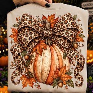 May include: A beige sweatshirt featuring a large graphic print of a pumpkin adorned with a prominent leopard print bow. The design includes orange autumn maple leaves, small brown acorns, and delicate berry sprigs surrounding the pumpkin.