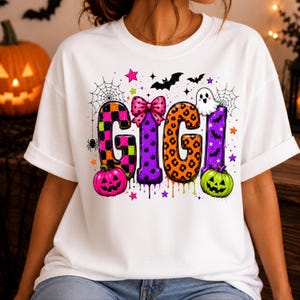 May include: A white t-shirt featuring the text GIGI in large, colorful, patterned letters. The design includes a pink polka dot bow, a white ghost, orange and green pumpkins, spider webs, bats, and small stars in a vibrant Halloween theme.