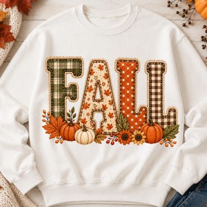 May include: A white crewneck sweatshirt featuring the word FALL in large, decorative letters. Each letter has a unique pattern: green plaid, maple leaf print, orange polka dots, and brown gingham. Orange pumpkins and autumn leaves decorate the base.