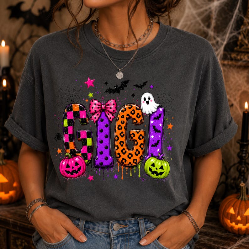 May include: A charcoal gray t-shirt featuring the text GIGI in large, colorful, patterned letters. The design includes a pink bow, a white ghost, orange and green jack-o'-lanterns, bats, stars, and spiderwebs in a vibrant Halloween theme.