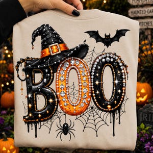 May include: A beige sweatshirt featuring a graphic print of the word BOO in black and orange, decorated with sparkling rhinestones. A black witch hat sits atop the B, with a flying bat, a spider, and a spiderweb design in the background.