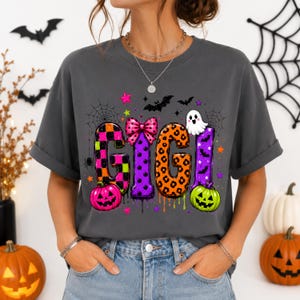 May include: A charcoal gray t-shirt featuring the word GIGI in large, colorful, patterned letters. The design includes a pink bow, a white ghost, orange and green pumpkins, spiderwebs, bats, and small stars in a vibrant Halloween theme.
