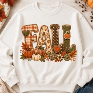 May include: A white crewneck sweatshirt featuring the word FALL in large, textured letters. The letters are decorated with plaid, floral, and polka dot patterns. Below the text are orange and white pumpkins, autumn leaves, and heart shapes.