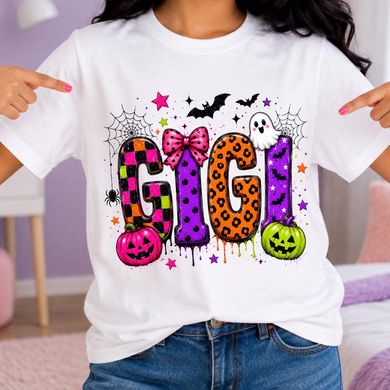 May include: A white t-shirt featuring a colorful graphic with the text GIGI in large letters. The letters are decorated with checkered, polka dot, and leopard patterns. The design includes pumpkins, a ghost, bats, spiders, and webs.