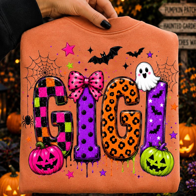 May include: An orange sweatshirt featuring the text GIGI in large, colorful letters. The letters are decorated with checkered, polka dot, and leopard patterns. The design includes a pink bow, a white ghost, bats, spiderwebs, and jack-o'-lanterns.