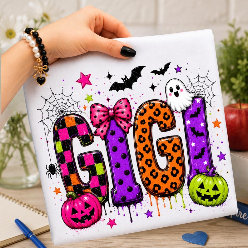 May include: A white fabric surface featuring the text GIGI in large, colorful letters. The letters are decorated with orange and black checkered, purple polka dot, and orange leopard patterns. Accents include a pink bow, a ghost, pumpkins, bats, and spiderwebs.