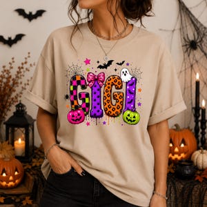 May include: A beige t-shirt featuring the word GIGI in large, colorful, patterned letters. The design includes a pink bow, a white ghost, a pink pumpkin, a green pumpkin, spiderwebs, bats, and small stars, all arranged in a festive Halloween theme.