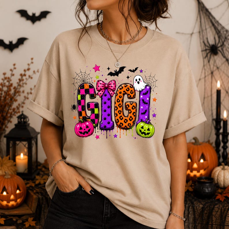 May include: A beige t-shirt featuring the word GIGI in large, colorful, patterned letters. The design includes a pink bow, a white ghost, a pink pumpkin, a green pumpkin, spiderwebs, bats, and small stars, all arranged in a festive Halloween theme.