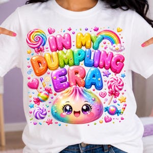 Può includere: Una t-shirt bianca con una grafica colorata che riporta il testo IN MY DUMPLING ERA in lettere a bolla 3D. Il design include un personaggio dumpling arcobaleno sorridente, vari lecca-lecca, caramelle, stelle e un arcobaleno dai colori vivaci.