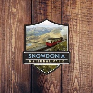 Może przedstawiać: Ilustracja w stylu vintage czerwonego pociągu jadącego w górę zbocza góry w Snowdonia National Park, Walii. Ilustracja ma kształt tarczy z tekstem "Snowdonia National Park" poniżej.