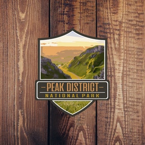 May include: A vintage-style national park badge with a brown background and a shield shape. The shield features a scenic view of a valley with green hills and a winding road. The text "Peak District National Park" is printed in brown letters on a white banner.