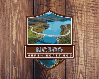 Può includere: Un adesivo marrone a forma di scudo con un'immagine blu e verde di un ponte su un corpo d'acqua. L'adesivo dice "NC500" e "NORTH COAST 500" in testo bianco.