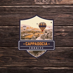 May include: A vintage-style travel sticker featuring a scenic view of Cappadocia, Turkey, with hot air balloons floating in the sky. The sticker has a shield shape with a brown border and the text "Cappadocia" and "TÜRKIYE" in white.