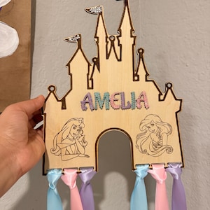 May include: A light-colored wooden castle-shaped wall hanging featuring the name AMELIA in glittery letters. The design includes engraved illustrations of two female characters and is decorated with small rhinestones on the spires. Six long satin ribbons in shades of light blue, pink, and lavender hang from the bottom edge.