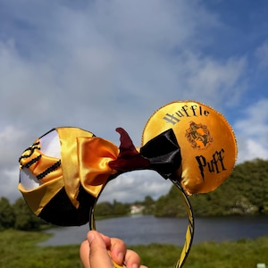 May include: A pair of headband ears featuring a yellow satin finish with black trim. The right ear displays the text Huffle Puff and a crest emblem. The left ear has black beaded accents. A central black and yellow bow sits between the ears.
