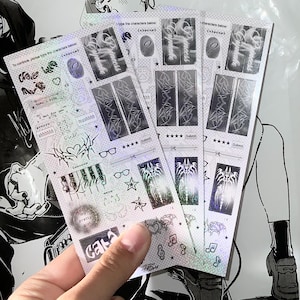 以下が含まれることがあります： 手には、いくつかのホログラフィックステッカーシートが握られています。ステッカーには、テキスト、星、ハート、抽象的なグラフィックなど、さまざまなデザインが施されています。シートは光沢のある虹色の仕上がりになっています。