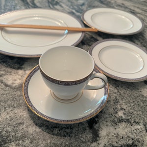 May include: A white teacup and saucer set with a blue and gold Greek key design. The set includes three oval plates and a large round plate, all with matching borders. The teacup is centered on the saucer, with the plates arranged around it.