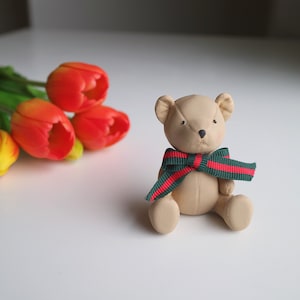 Könnte beinhalten: Ein kleiner, brauner Teddybär mit einer grün-roten Schleife. Der Bär sitzt auf einer weißen Oberfläche.