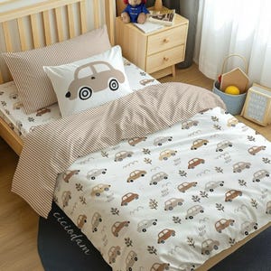 Può includere: Un set di biancheria da letto con copripiumino e federa bianchi con un motivo ripetuto di piccole auto marroni e grigie, alberi e il testo BEEP-BEEP. Una federa presenta una grande grafica di un'auto beige. Il tessuto ha strisce marroni.