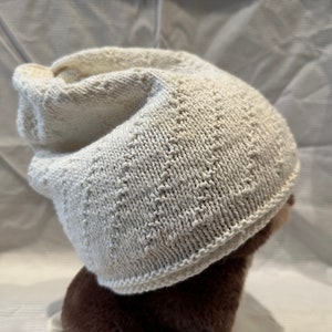 May include: Off-white knitted beanie hat. The hat features a textured pattern and a folded brim. The hat is made of a soft, warm material, perfect for cold weather. The hat is a neutral color, making it versatile for any outfit.