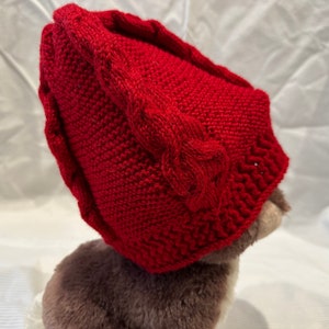 May include: A red knitted hat with a cable knit design. The hat is made of soft yarn and has a pointed crown.
