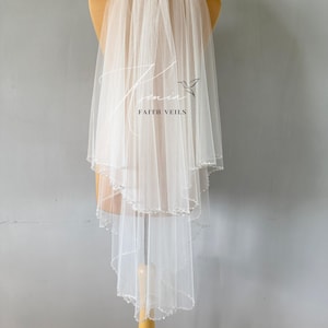 May include: A white, layered bridal veil with a scalloped edge and beaded trim. The veil is displayed on a mannequin and features the text "Tamin Faith Veils". The veil is made of a sheer, lightweight fabric.
