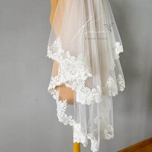 May include: A two-tiered white bridal veil with delicate lace trim. The veil is displayed on a mannequin and features the text "Tsonia Faith Veils" in a script font.