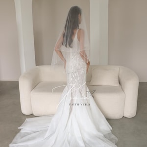 May include: A bride in a white wedding dress with a long train and a veil. The dress has a fitted bodice and a flared skirt. The veil is sheer and covers the bride's head and shoulders. The text "FAITH VEILS" is visible.
