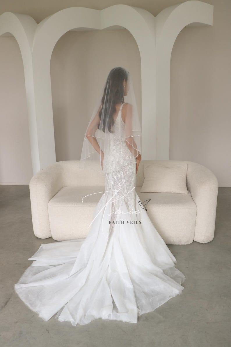 May include: A bride in a white wedding dress with a long train and a veil. The dress has a fitted bodice and a flared skirt. The veil is sheer and covers the bride's head and shoulders. The text "FAITH VEILS" is visible.