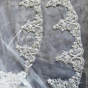 May include: Close-up of a white bridal veil with intricate, ornate lace detailing. The lace features floral and swirling patterns, embellished with small, delicate beads. The veil is set against a soft gray background.