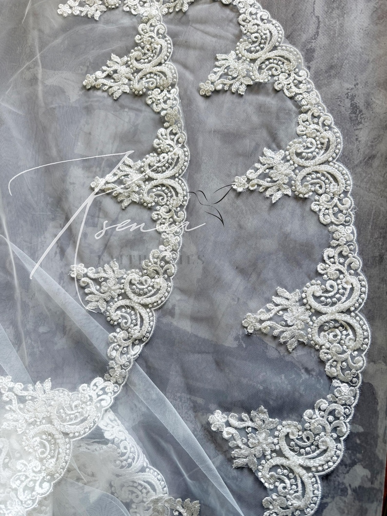 May include: Close-up of a white bridal veil with intricate, ornate lace detailing. The lace features floral and swirling patterns, embellished with small, delicate beads. The veil is set against a soft gray background.