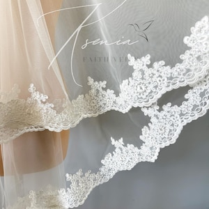 May include: Close-up of a white bridal veil with delicate floral lace trim. The veil is sheer and layered, with the text "Sennia Faith Veils" in a script font. The lace trim adds a touch of elegance to the bridal accessory.
