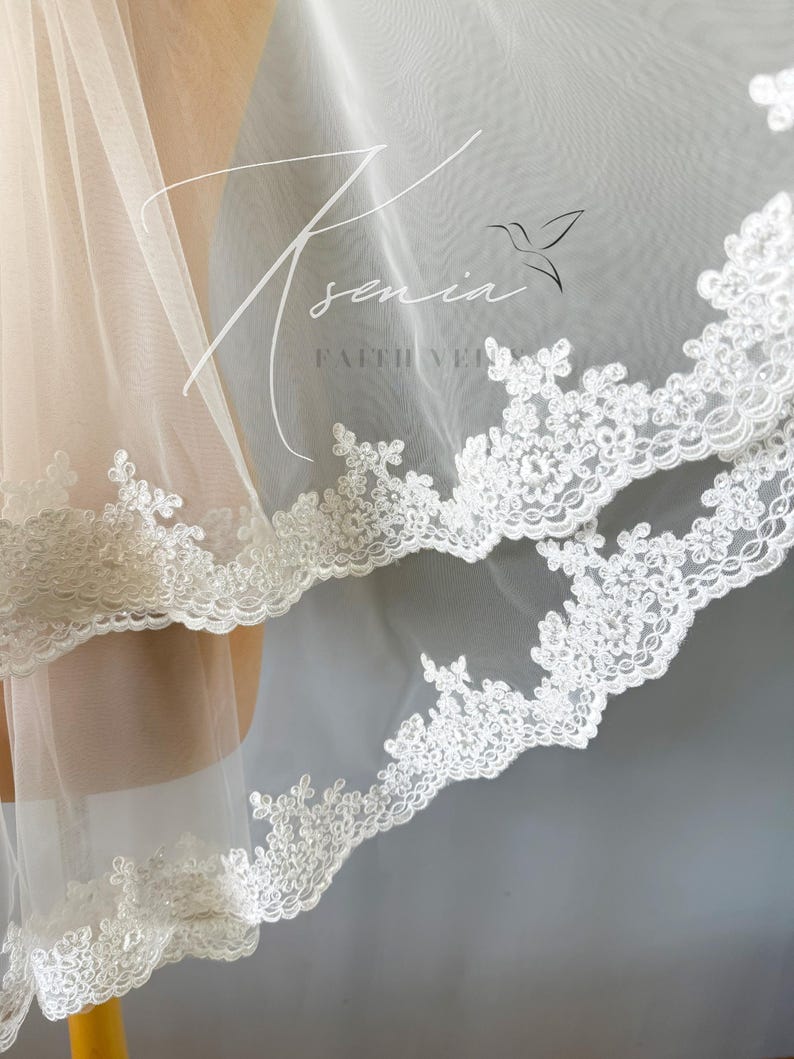 May include: Close-up of a white bridal veil with delicate floral lace trim. The veil is sheer and layered, with the text "Sennia Faith Veils" in a script font. The lace trim adds a touch of elegance to the bridal accessory.