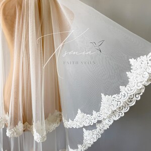 May include: A sheer, ivory-colored bridal veil with a delicate floral lace trim. The veil is draped over a mannequin, showcasing its flowing fabric and intricate design. The text "Tsenia Faith Veils" is visible.