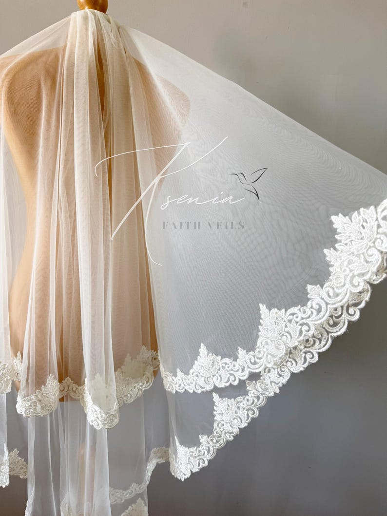 May include: A sheer, ivory-colored bridal veil with a delicate floral lace trim. The veil is draped over a mannequin, showcasing its flowing fabric and intricate design. The text "Tsenia Faith Veils" is visible.