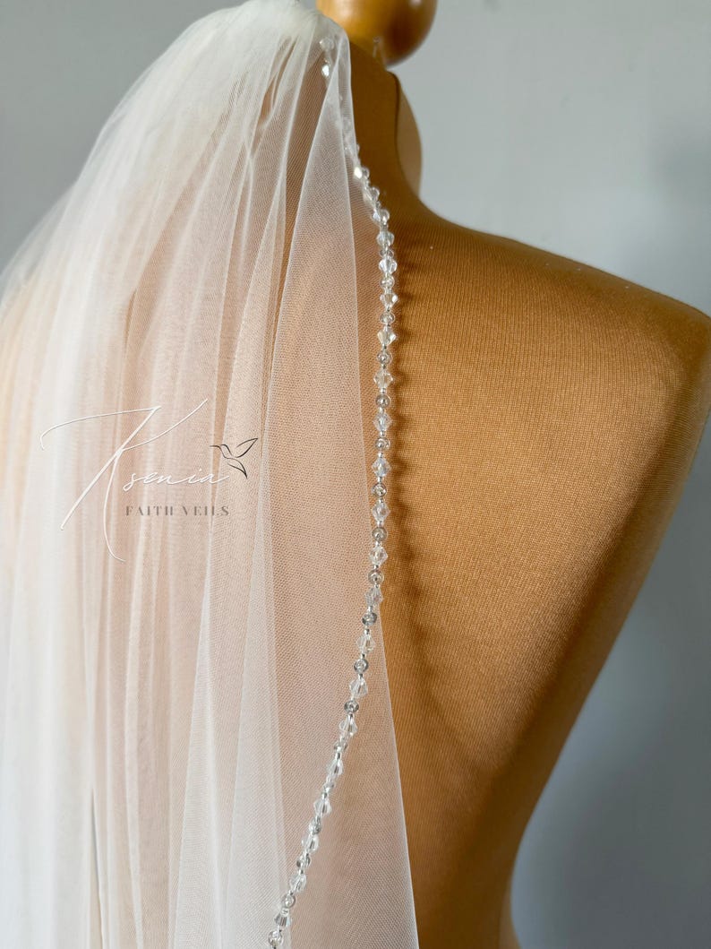 May include: A sheer, white bridal veil with a delicate beaded edge. The veil is draped over a mannequin, showcasing its flowing fabric and intricate crystal detailing. The text "Faith Veils" is visible on the veil.