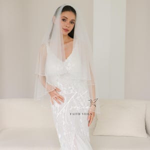 May include: A bride wearing a white wedding dress and veil. The dress is embellished with intricate beadwork. The veil is a sheer, two-tiered design. The text "FAITH VEILS" is visible on the dress.