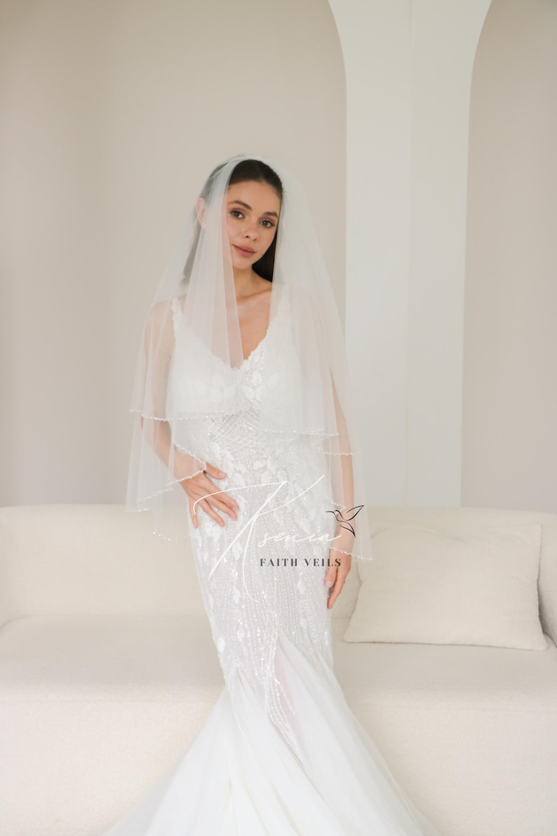 May include: A bride wearing a white wedding dress and veil. The dress is embellished with intricate beadwork. The veil is a sheer, two-tiered design. The text "FAITH VEILS" is visible on the dress.