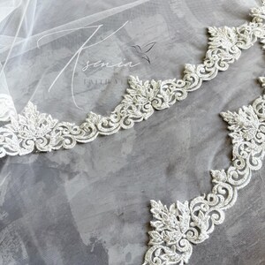 May include: A close-up of a white bridal veil with intricate floral lace edging. The delicate lace trim features detailed leaf and scroll patterns. The veil is sheer, with the text "Ksenia Faith Veils" and a hummingbird logo.