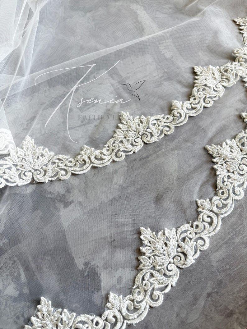 May include: A close-up of a white bridal veil with intricate floral lace edging. The delicate lace trim features detailed leaf and scroll patterns. The veil is sheer, with the text "Ksenia Faith Veils" and a hummingbird logo.