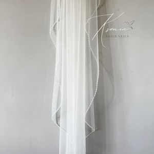 May include: A sheer white bridal veil with a delicate beaded edge, hanging against a gray wall. The veil is long and flowing, with a subtle shimmer. The text "Asenia Faith Veils" is visible in the upper right corner.