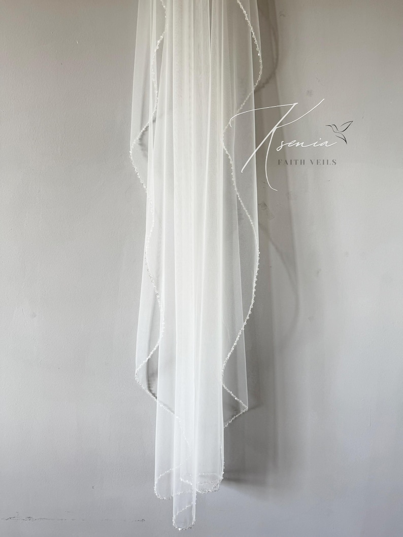 May include: A sheer white bridal veil with a delicate beaded edge, hanging against a gray wall. The veil is long and flowing, with a subtle shimmer. The text "Asenia Faith Veils" is visible in the upper right corner.