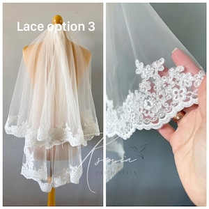 May include: A two-tiered white bridal veil with delicate floral lace trim. The veil is displayed on a mannequin, showcasing the soft, flowing fabric and intricate lace detailing. The text "Lace option 3" is visible.